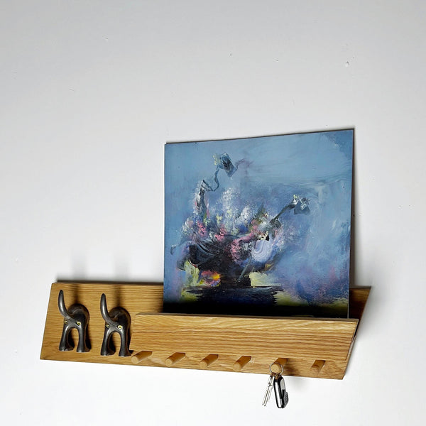 May include: A light oak shelf with two black elephant hooks and several pegs. A framed painting with a blue background and floral imagery is displayed on the shelf. Keys hang from the bottom of the shelf.