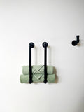 May include: Black metal towel rack with two bars and a single hook mounted on a white wall. Two green towels are folded and hanging on the bars.